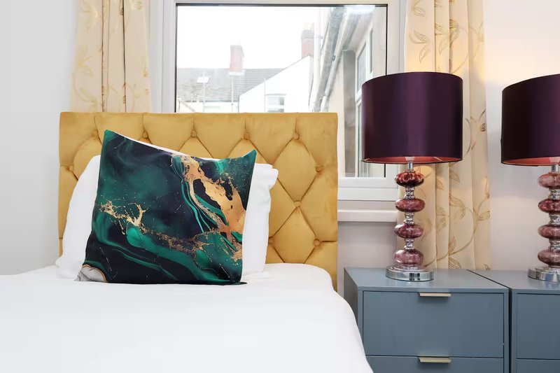 Modern bedroom with gold upholstered headboard, white bedding, decorative pillows, and burgundy table lamps