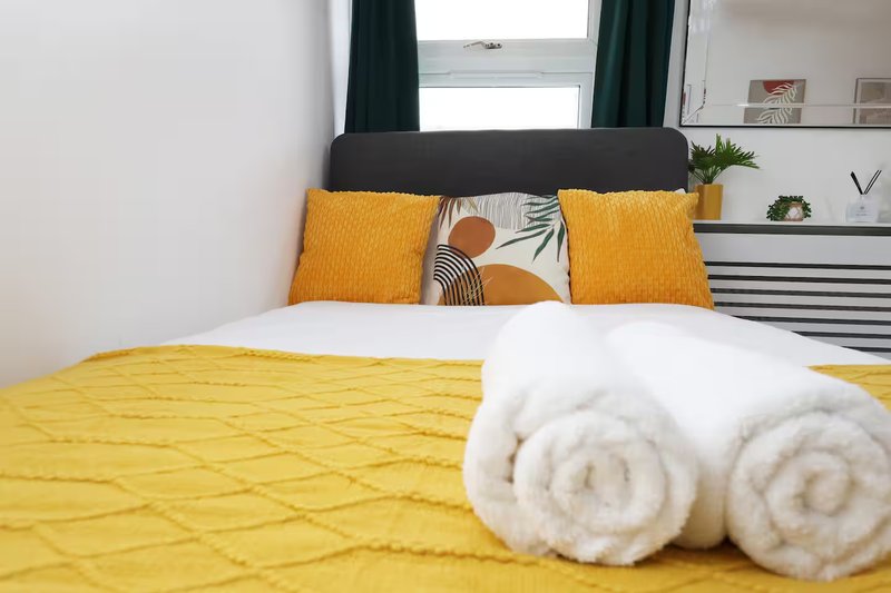 Bedroom with yellow quilted bedding, orange and patterned cushions, white rolled towels, and window with dark curtains