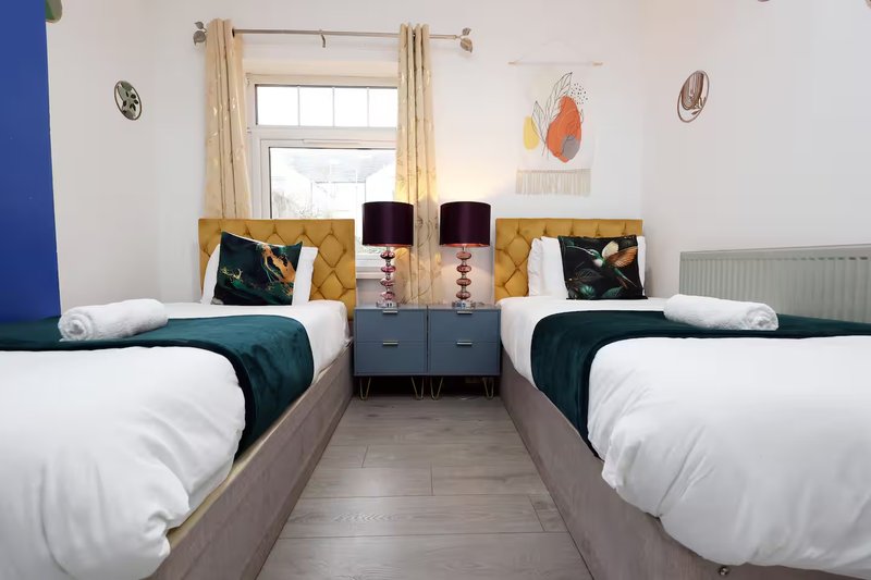 Twin bedroom with two beds, bedside table, lamps, window with curtains, and wall art