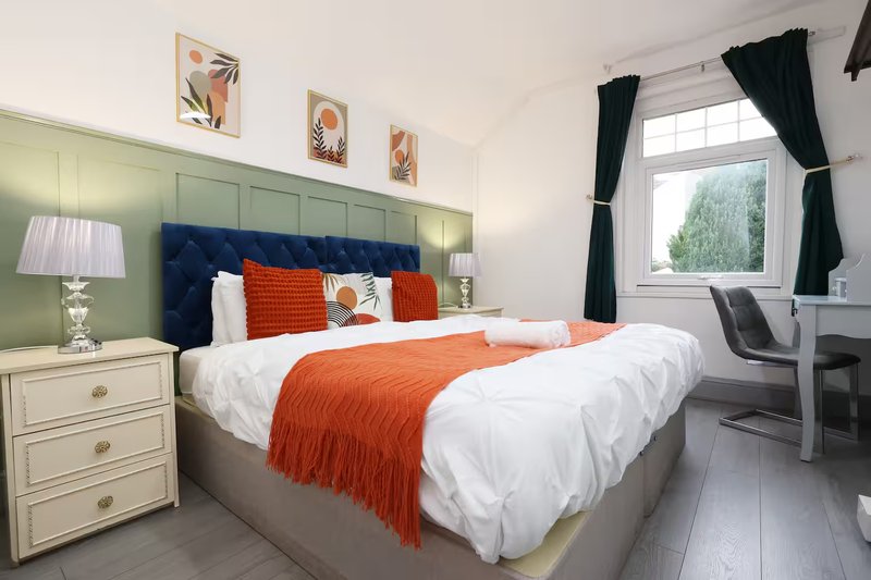 Bedroom with double bed, orange throw, blue headboard, green panelled wall and white nightstand