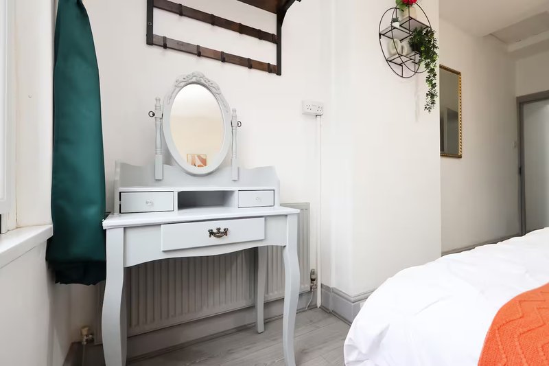 Bedroom with white vanity mirror, desk, hanging plants, and bed with orange throw