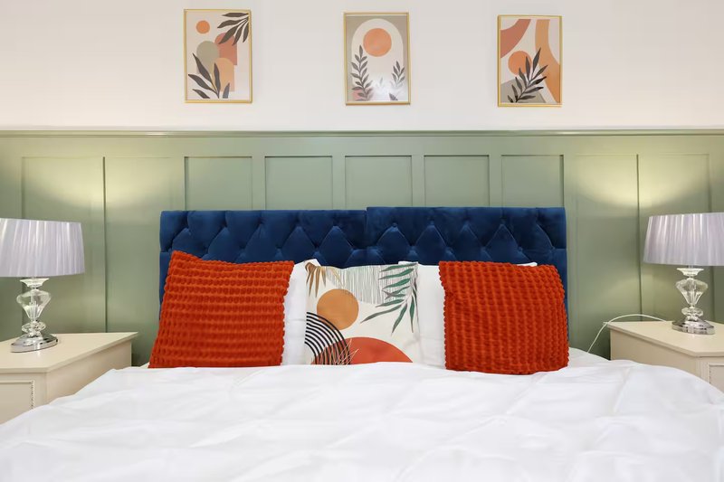 Modern bedroom with blue upholstered headboard, white bedding, red and white decorative pillows, bedside lamps, and botanical wall art