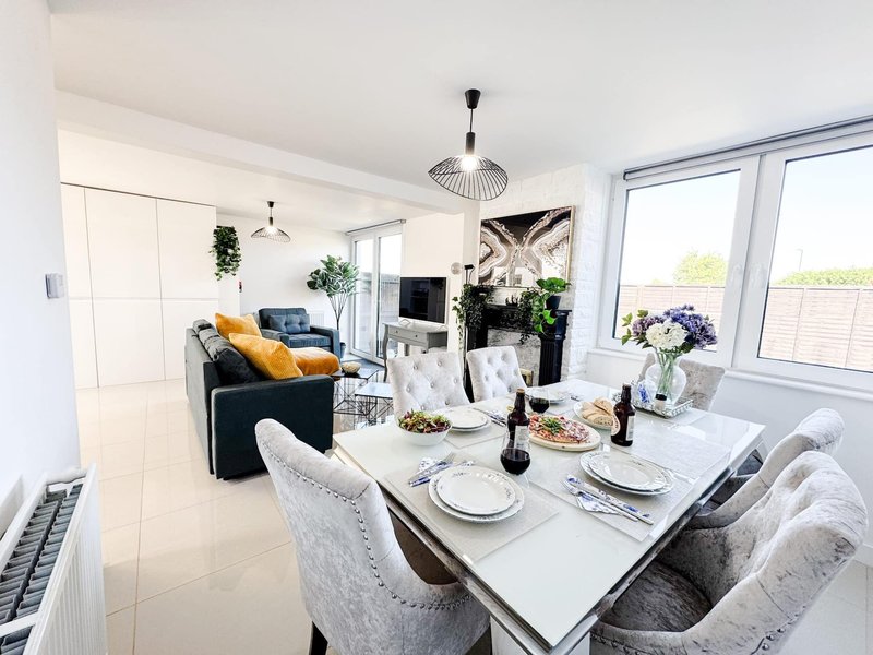 Open-plan dining and living room with white table, upholstered chairs, and grey sofa with yellow cushions