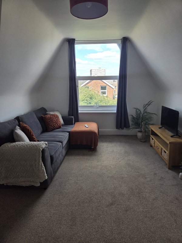 Bright attic bedroom with grey sofa, brown ottoman, window overlooking brick house, and wooden TV unit.