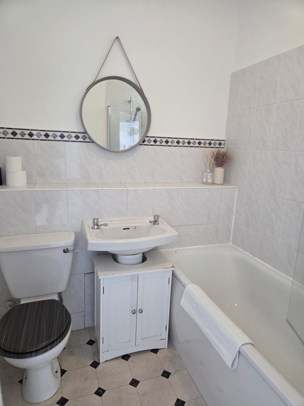 Compact bathroom with toilet, pedestal sink, circular mirror, bathtub and black-and-white tile flooring