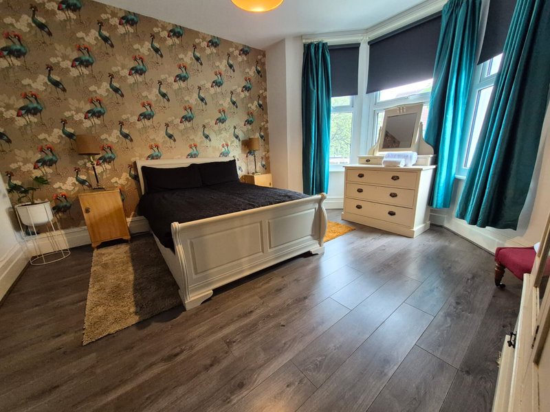 Spacious bedroom with double bed, wooden flooring, patterned wallpaper and turquoise curtains