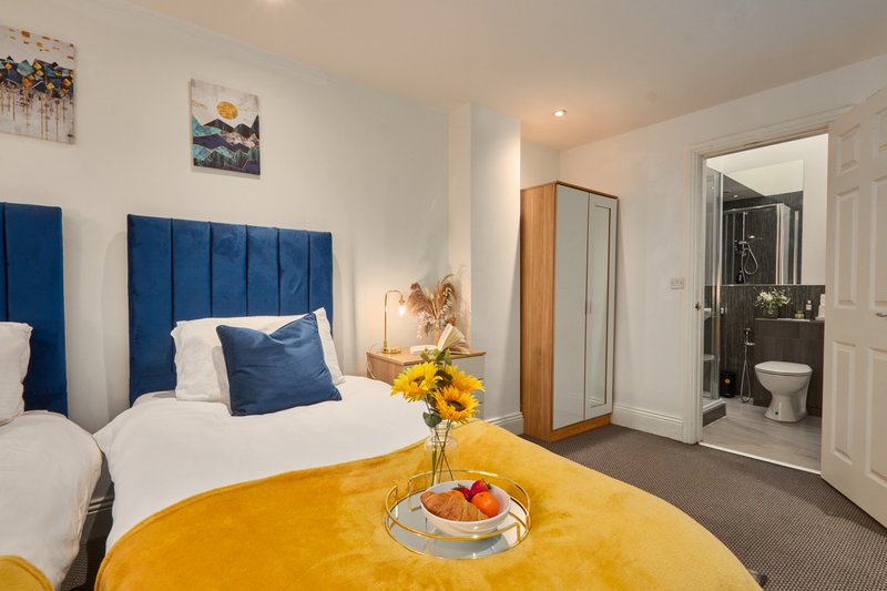 Bright bedroom with blue upholstered headboard, white bedding, yellow ottoman, and ensuite bathroom visible