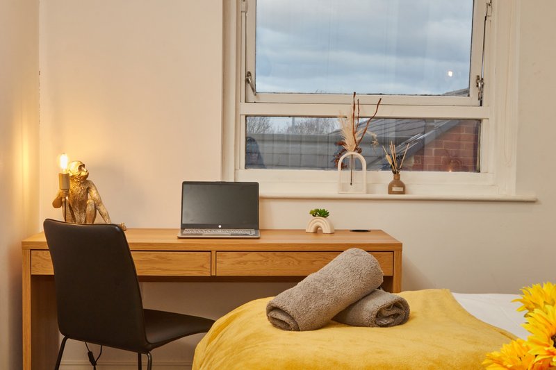 Bright bedroom with wooden desk, laptop, chair, and yellow bedding with rolled towels