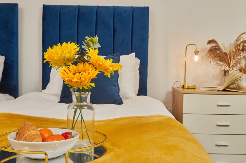 Modern bedroom with blue upholstered headboard, yellow flowers in vase, and fruit bowl on bed