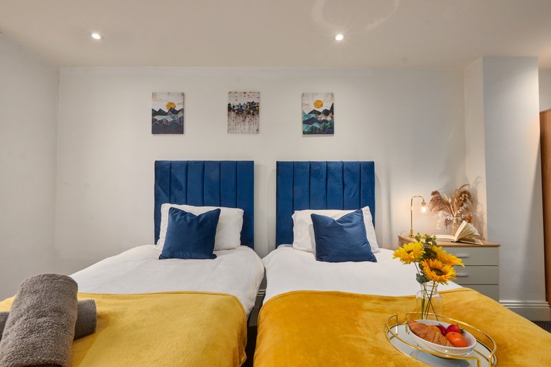 Twin bedroom with blue upholstered headboards, yellow bedding, framed wall art, and side table with flowers
