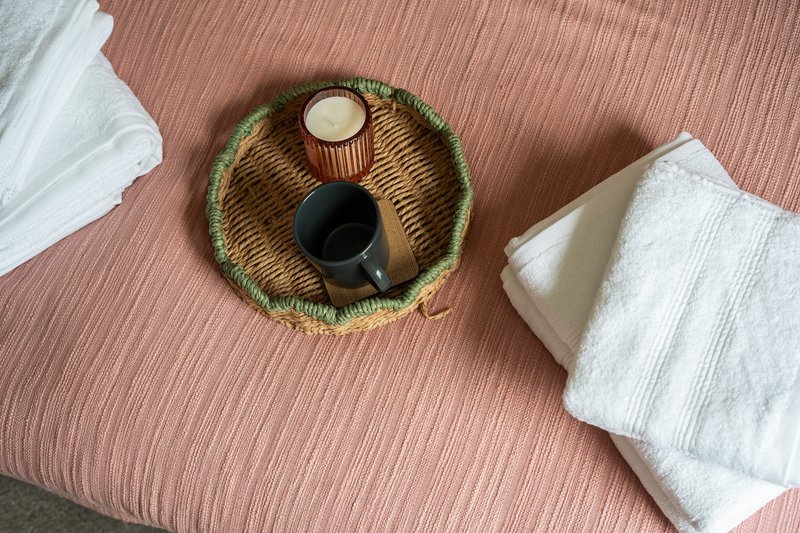Woven basket with candle and brush on pink textured fabric, white towels nearby