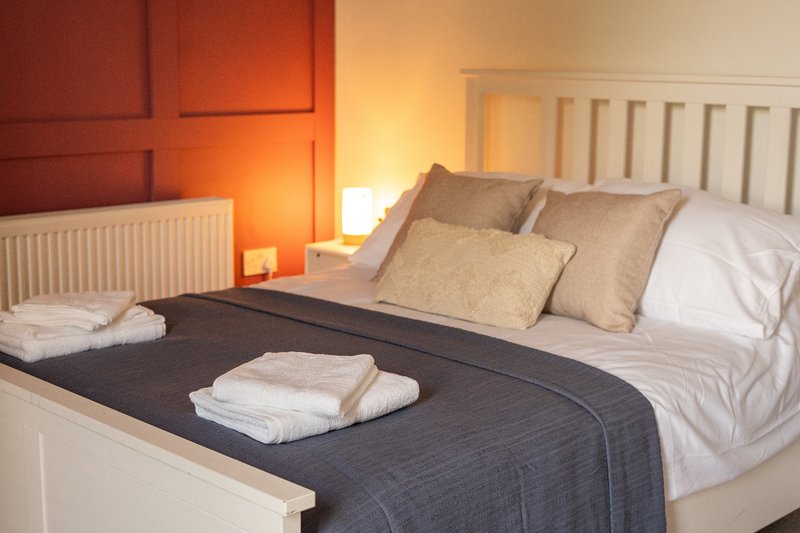 Bedroom with double bed, white headboard, warm lighting, and folded towels on bedding