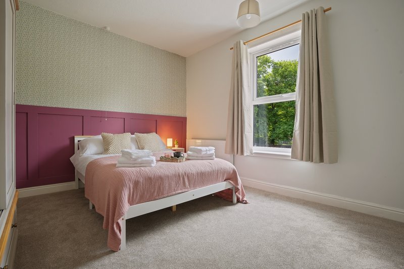 Bedroom with pink upholstered headboard, white bed frame, pink bedding, and window with garden view