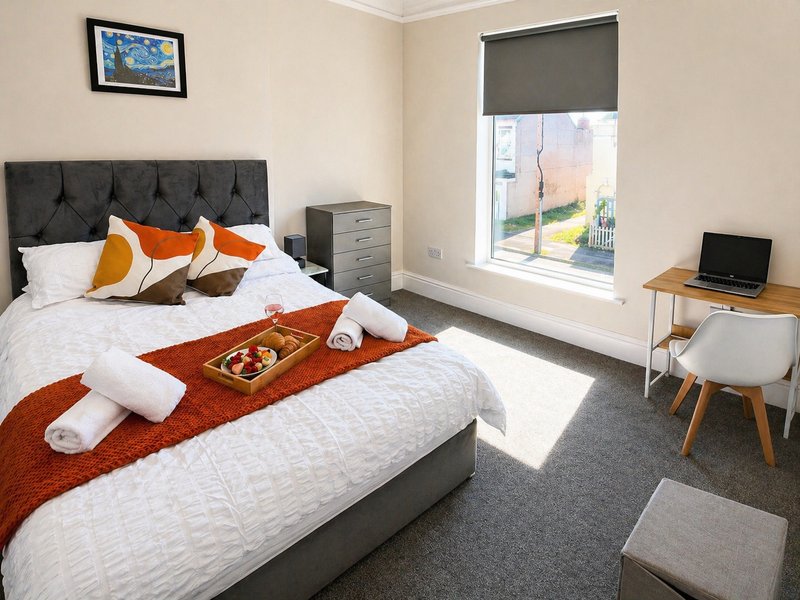 Bright bedroom with double bed, orange throw, decorative pillows, desk with laptop and window view