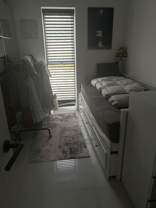 Narrow bedroom with single bed, venetian blinds window, white walls and polished floor