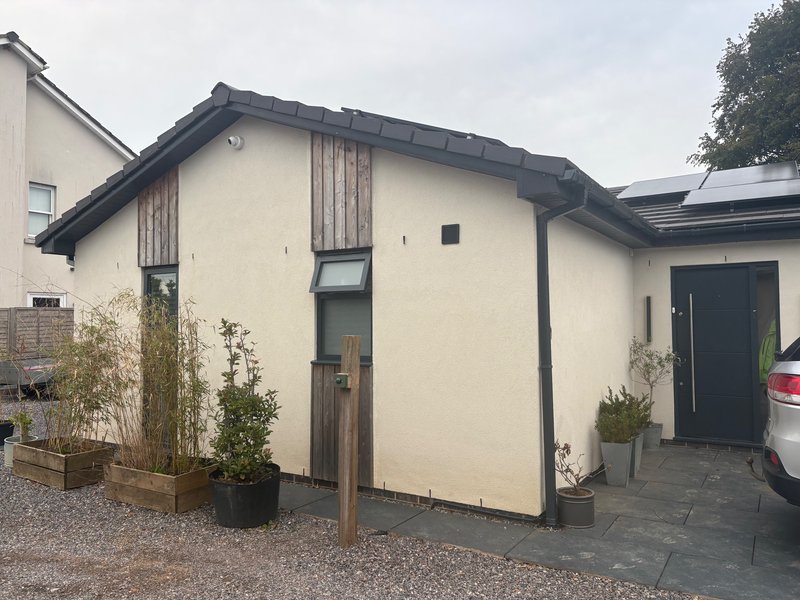 Modern single-story residential building with cream walls, dark roof, wooden entrance door and potted plants