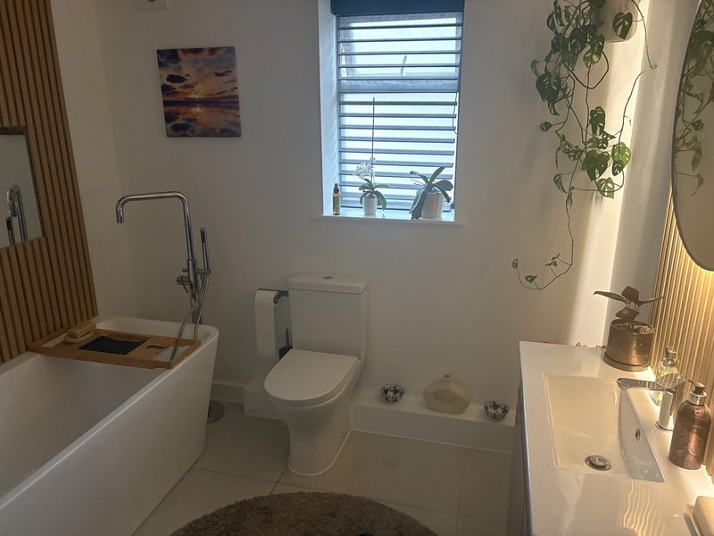 Modern bathroom with white bathtub, toilet, dual sinks, window with blinds and wall art