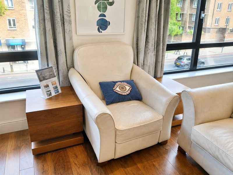 Cream upholstered armchair with wooden side table, decorative pillow, and large windows overlooking street