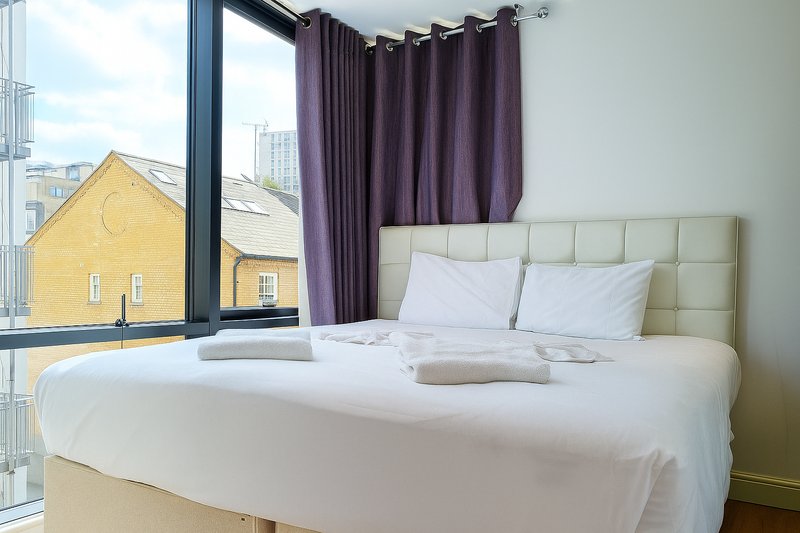 Modern bedroom with double bed, purple curtains, large window overlooking city street