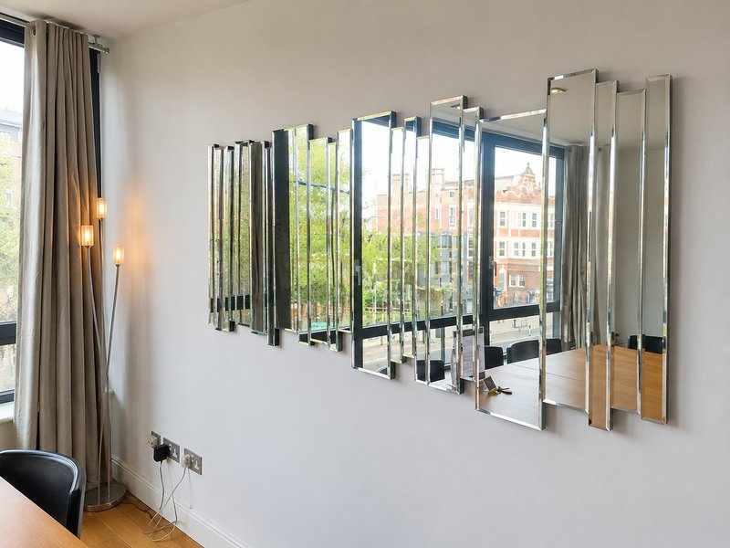 Modern bedroom with mirrored wall art, cream curtains, and city views through windows