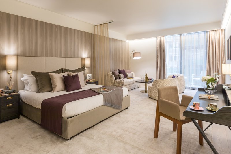 Spacious hotel bedroom with king bed, seating area, desk, and city views from floor-to-ceiling windows