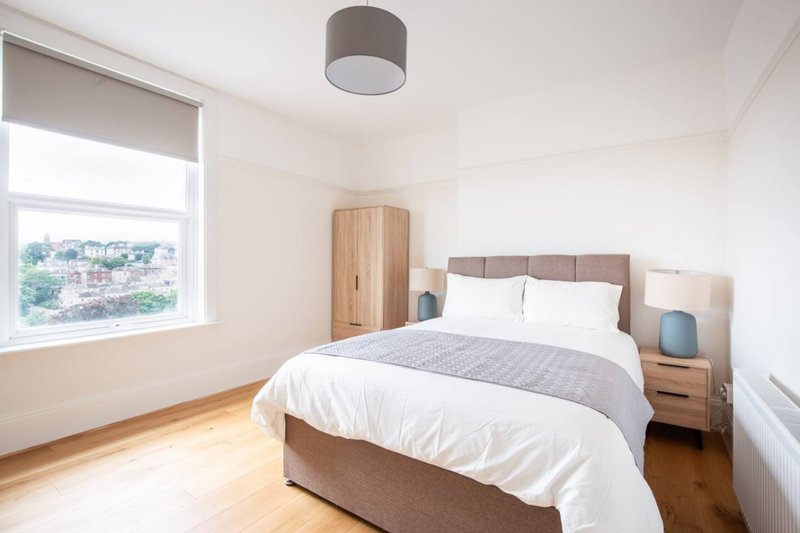 Bright bedroom with double bed, wooden wardrobe, and large window overlooking cityscape