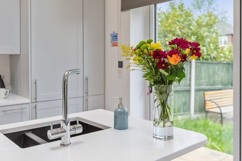 Modern white kitchen with island counter, chrome tap, fresh flowers in vase, and garden view