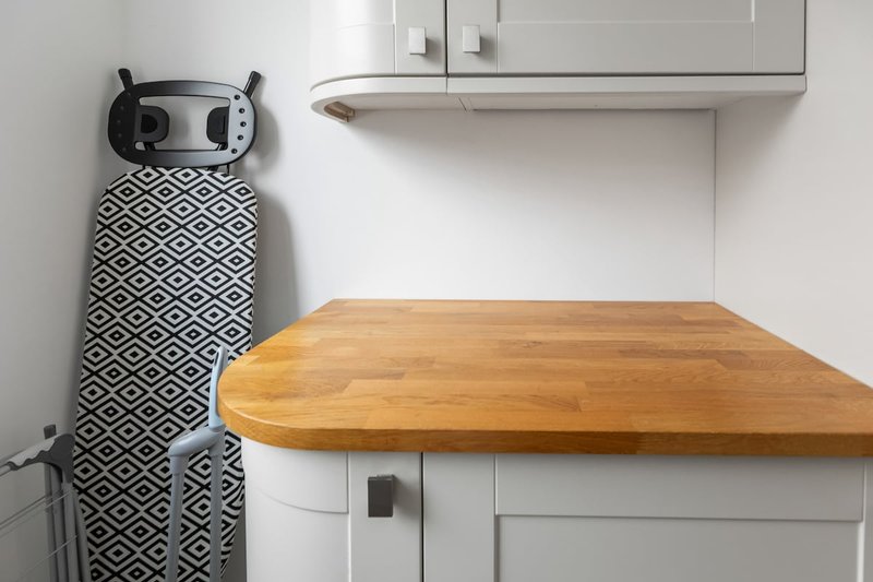 Kitchen counter with wooden surface, white cabinetry, and ironing board leaning against wall