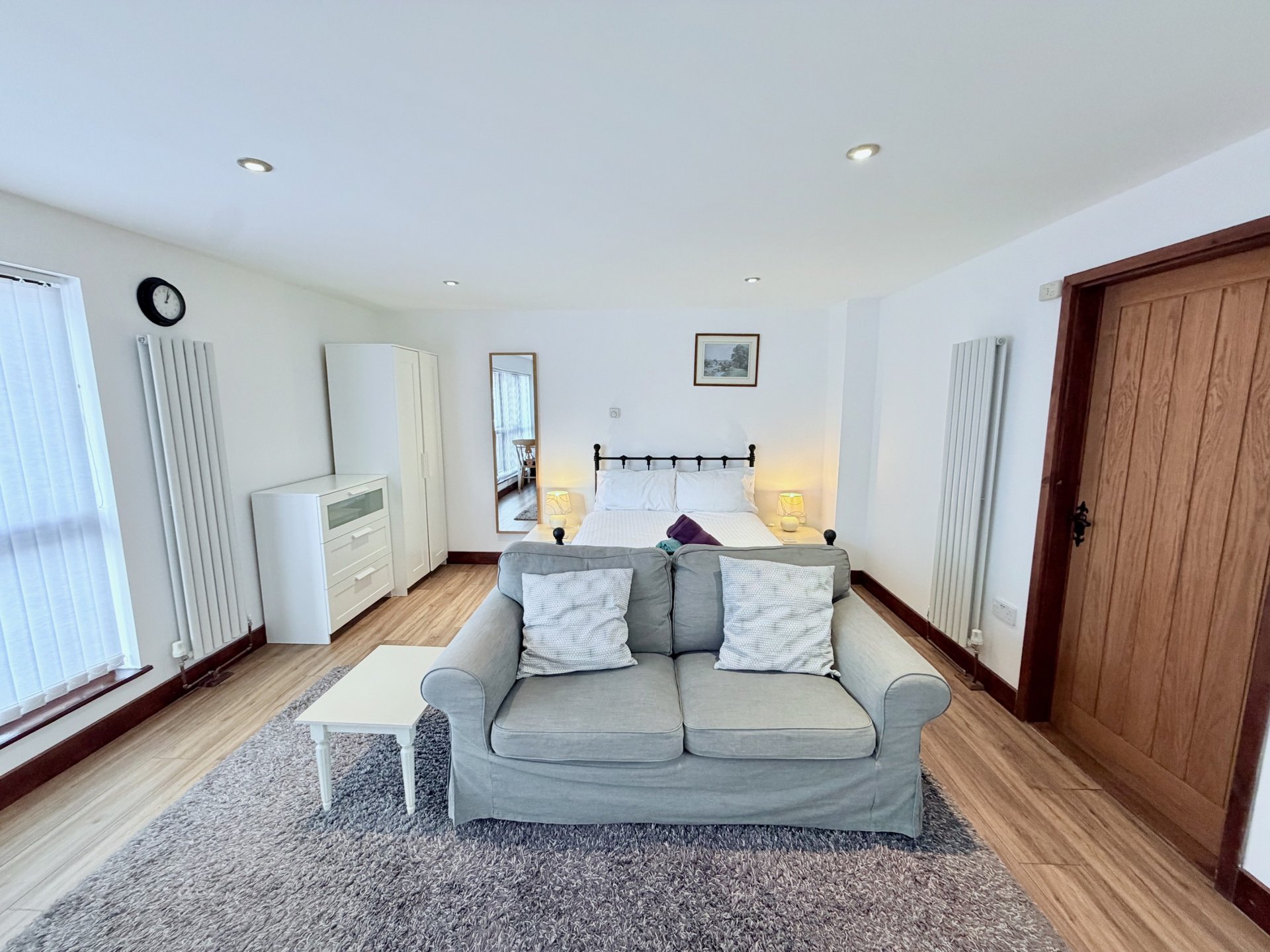 Spacious bedroom with gray sofa, double bed, wooden flooring and white walls