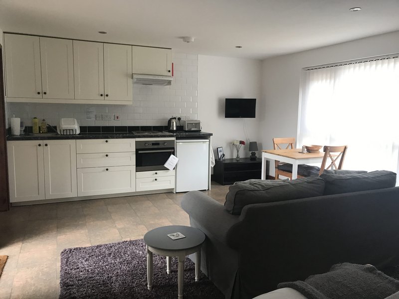 Open-plan kitchen and living area with cream cabinets, black sofa, dining table and wall-mounted TV
