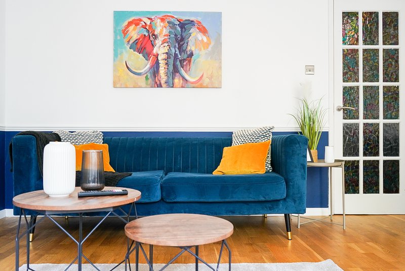 Modern living room with blue velvet sofa, wooden coffee tables, colorful wall art and orange cushions