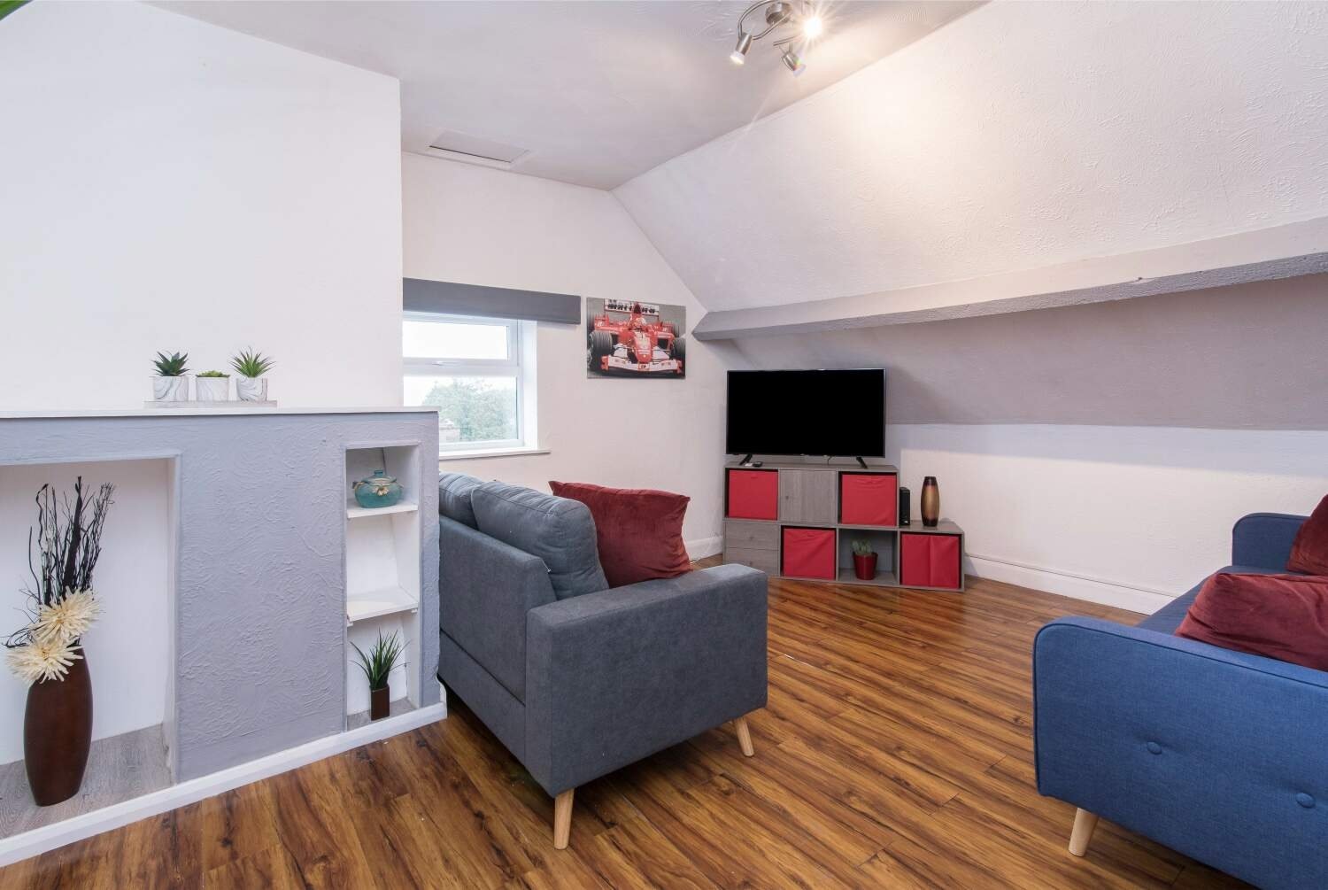 Attic living room with gray armchair, blue sofa, red cushions, wooden flooring and TV.