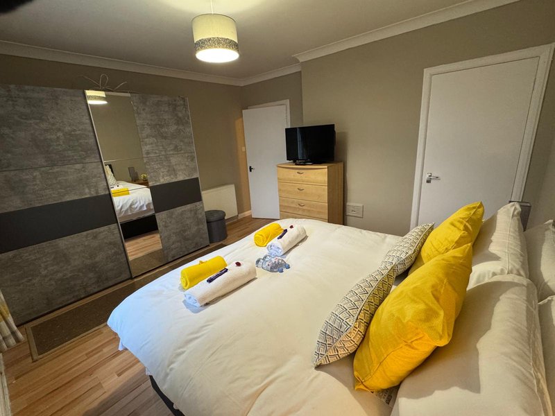 Spacious bedroom with double bed, yellow pillows, wooden dresser, mirror, and wall-mounted TV