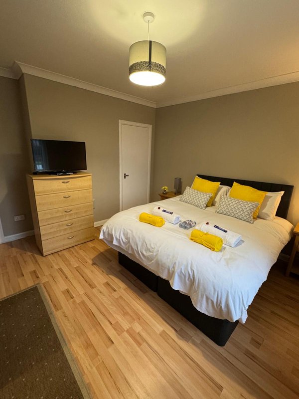 Bright bedroom with double bed, yellow cushions, wooden dresser, TV, and pendant ceiling light