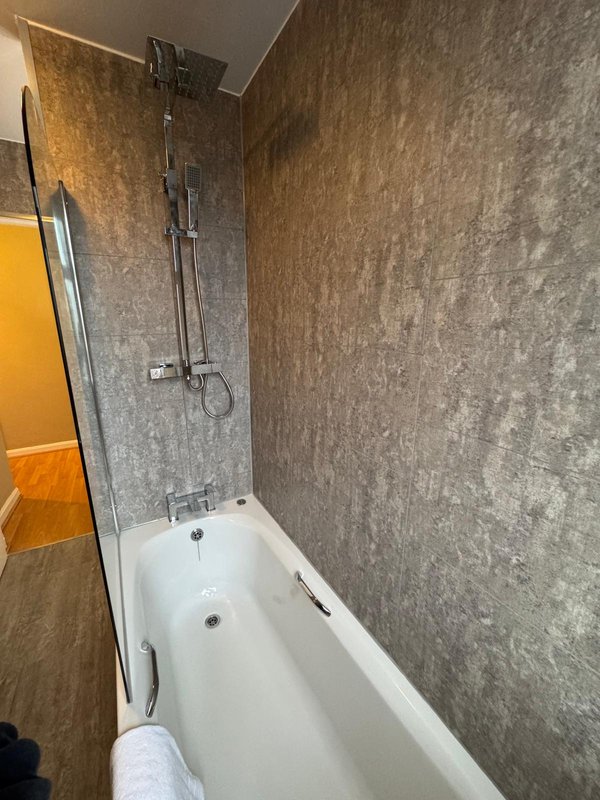 Bathroom with white bathtub, grey tile walls, overhead shower and glass panel