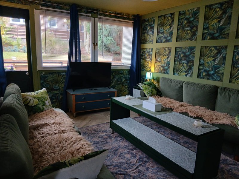 Living room with green upholstered seating, TV, botanical wall art, and large windows