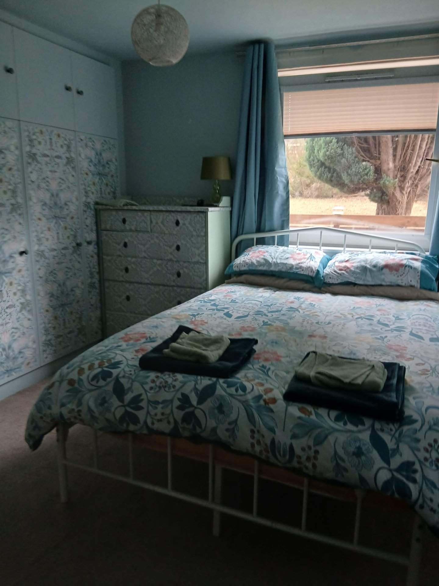 Bedroom with double bed, grey dresser, blue walls, window with blinds and curtains