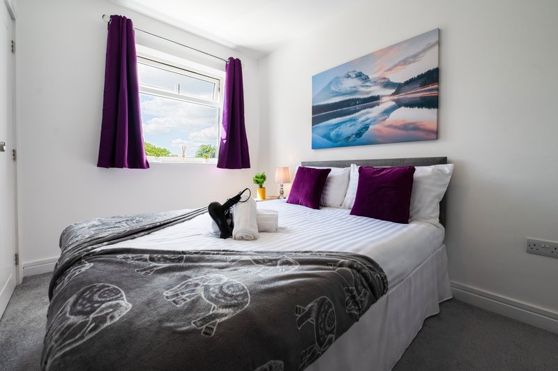 Modern bedroom with double bed, purple curtains, landscape artwork, and natural light from window