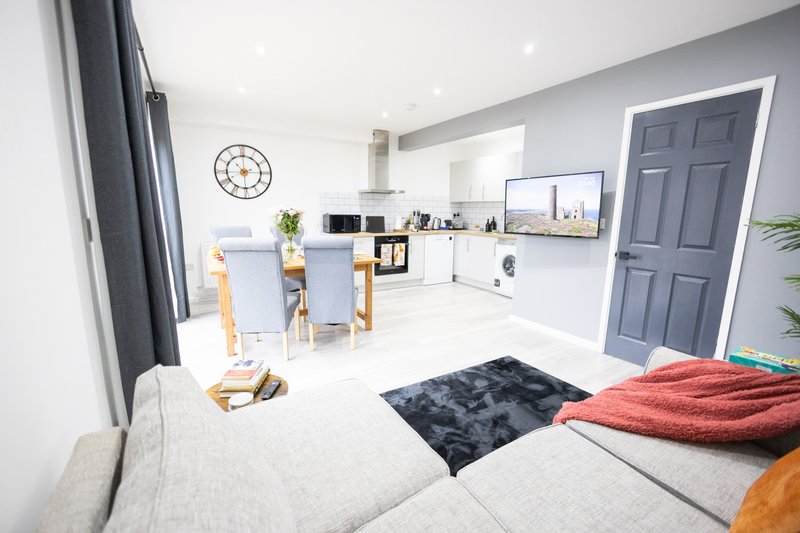 Open-plan living area with kitchen, dining table, sofa, and bedroom space with clock on wall