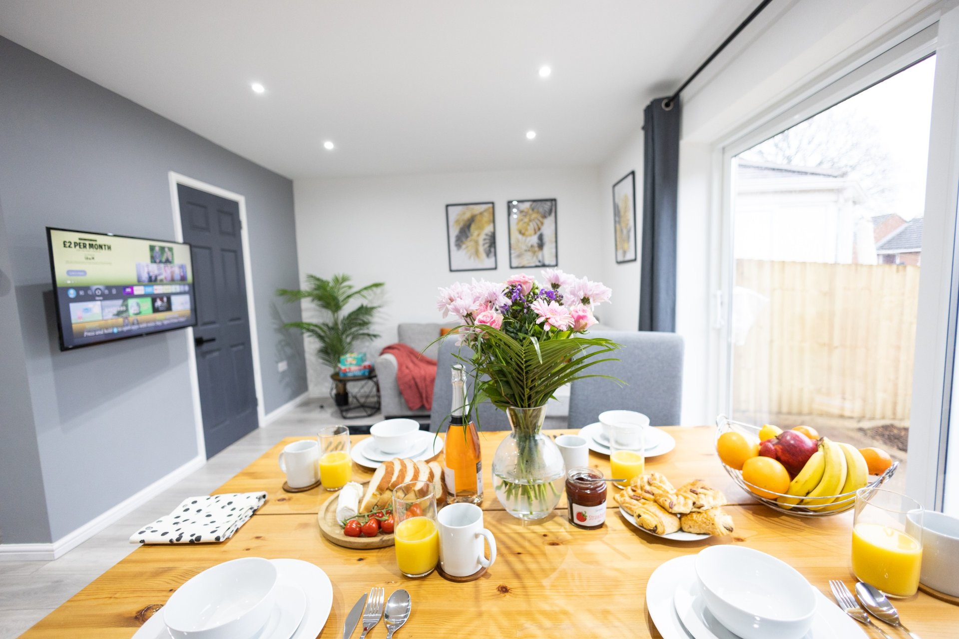 Modern dining room with wooden table set for breakfast, fresh flowers, and garden view through patio doors