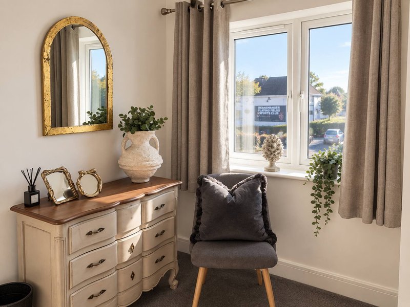 Bedroom corner with wooden dresser, gold-framed mirror, black chair, and window with garden view
