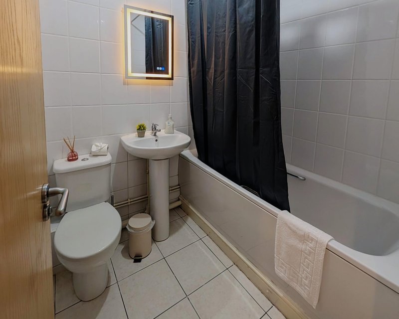 Clean bathroom with toilet, pedestal sink, bathtub, black shower curtain and illuminated mirror