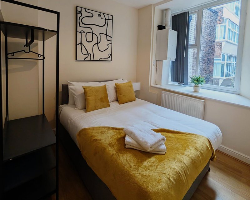 Modern bedroom with double bed, gold velvet bedding, yellow cushions, wall art, and city view from window
