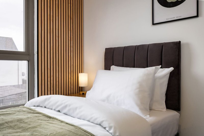 Modern bedroom with black upholstered headboard, white bedding, wooden slat wall paneling, and city view
