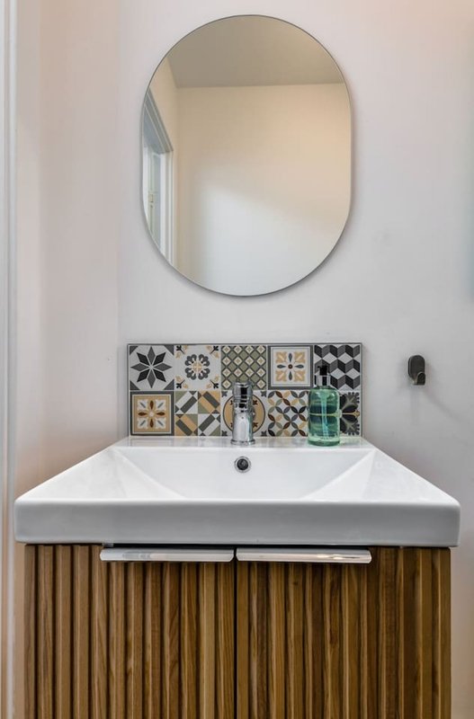 Modern bathroom with white sink, wooden vanity, patterned tile backsplash and oval mirror