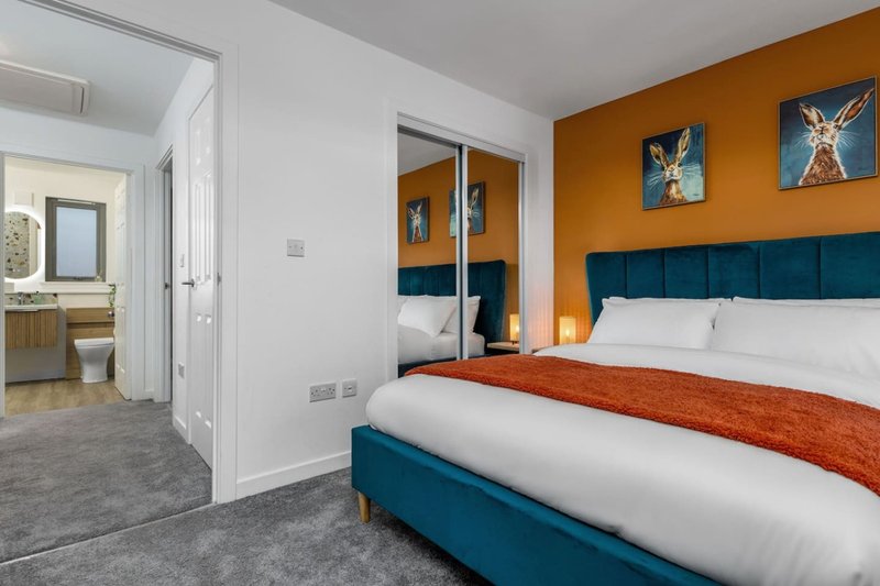 Modern bedroom with teal upholstered bed, orange accent wall, ensuite bathroom visible through doorway
