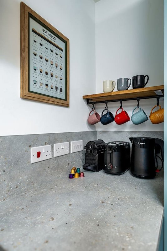 Kitchen counter with coffee maker, mugs on shelf, and framed artwork on wall