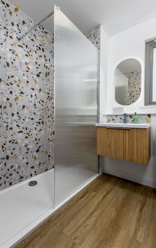 Modern bathroom with terrazzo tile walls, wooden vanity, frosted glass shower enclosure and round mirror