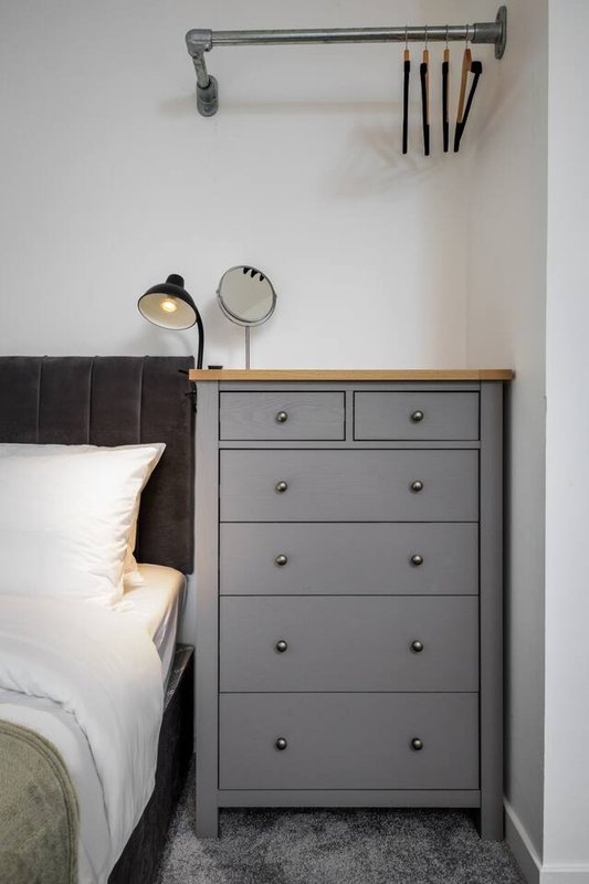 Bedroom corner with gray chest of drawers, bedside lamp, and industrial pipe shelving