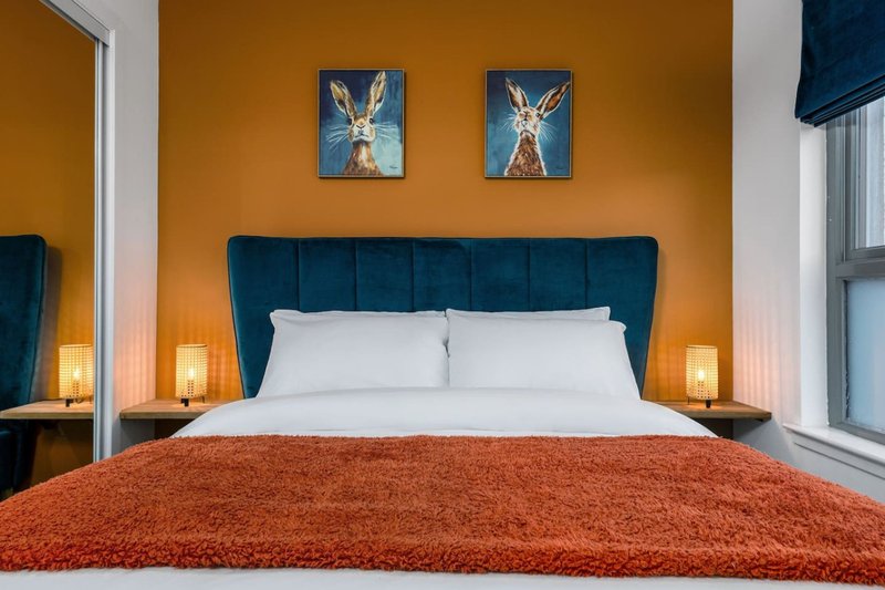 Bedroom with teal upholstered headboard, white pillows, orange throw blanket, and framed wall art